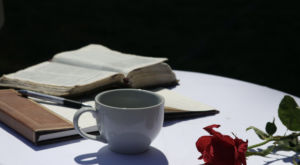 An open-faced Bible, Coffee cup, and rose on a white tablecloth.